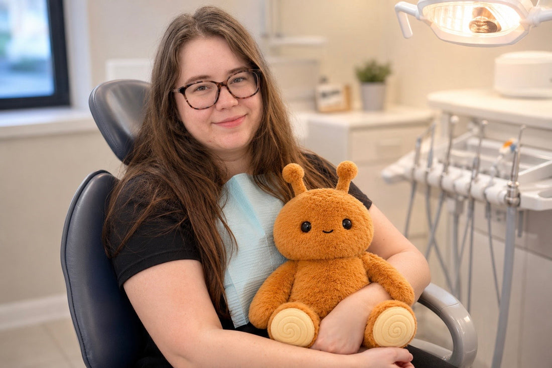 Adult woman sitting in a dentist chair holding Pebble, a brown Feelikins sensory plush toy, in a calm dental office setting.