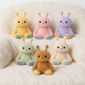 Six cute sensory plush toys in pastel colors arranged on a soft cream-colored plush chair. The are yellow, pink, purple, green, brown, and cream, each with small antennae, black embroidered eyes, and pastel-colored feet.