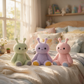 Three cute plush self care sensory toys in pastel green, lavender, and pink are sitting on a cozy bed with cream-colored bedding in a sunlit bedroom.