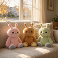 Three cute sensory toy emotional support plush toys in pastel pink, mustard yellow, and mint green sitting on a beige sofa in a sunlit living room.