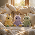 Three pastel sensory plush toys sit side by side on a soft cream blanket with warm fairy lights in the background. A small pillow fort surrounds them. 