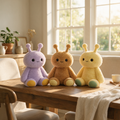 Three cute sensory fidget plush toys sitting on a wooden dining table in a warm, sunlit room. The toys have long antennae and simple embroidered faces.