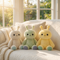 Three cute plush toys in pastel colors of  cream, green, and yellow, sitting side by side on a cozy cream-colored sofa in a sunlit room.