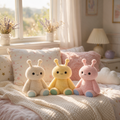 Three cute plush toys in cream, yellow, and pastel pink are arranged on a cozy bed with pink and white pillows in a warm, sunlit bedroom.