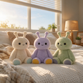 Three cute plush toys in pastel colors (cream, lavender, and green) sitting together on a cozy bed in a warm, sunlit bedroom.