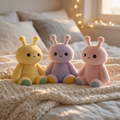 Three cute plush emotional support toys in pastel yellow, lavender, and pink, sitting together on a cozy cream knit blanket on a bed with warm fairy lights in the background.